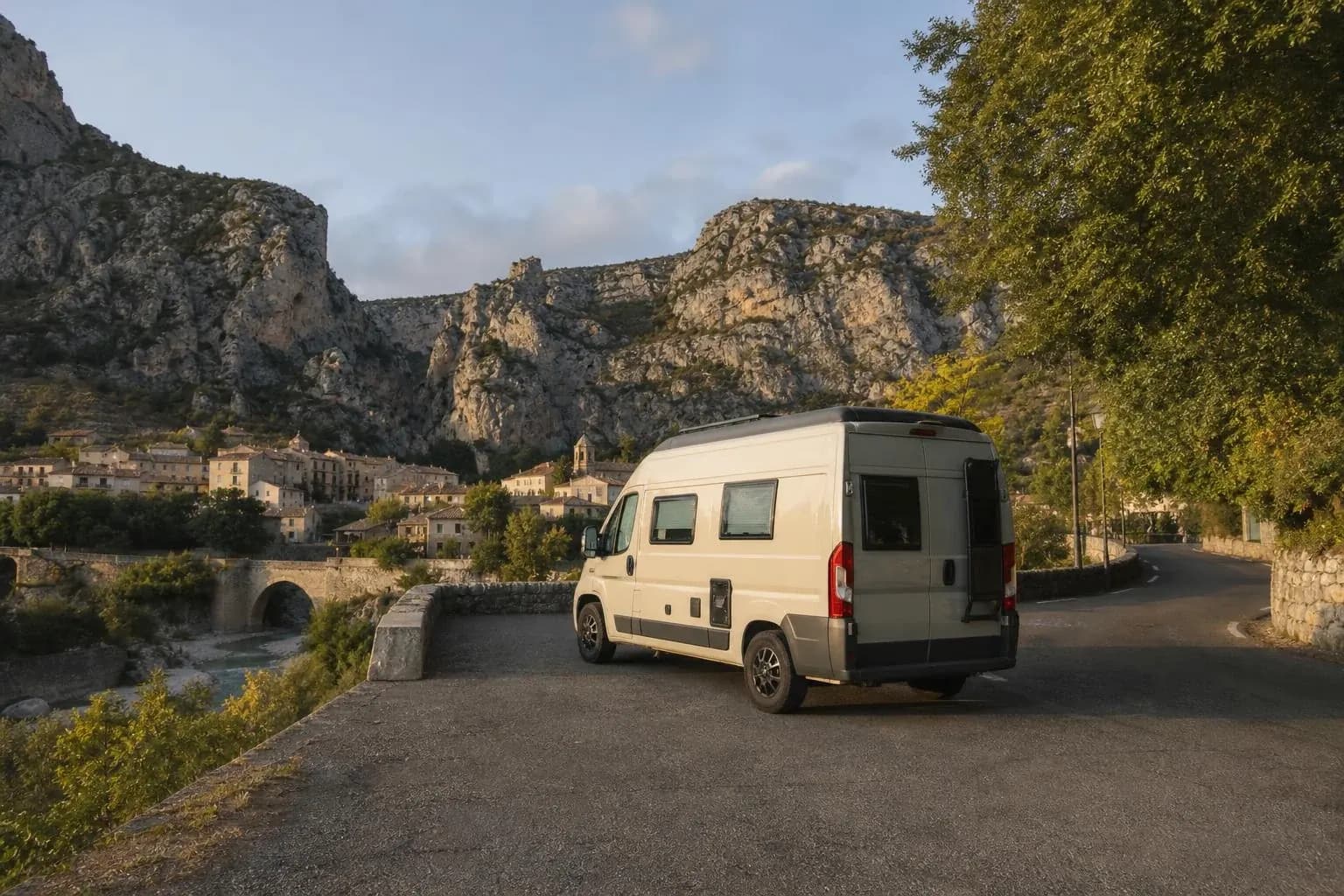 Van aménagé blanc vu de trois-quarts arrière, garé en surplomb d'un vieux pont de pierre franchissant des gorges, face à un village accroché à la falaise, au petit matin